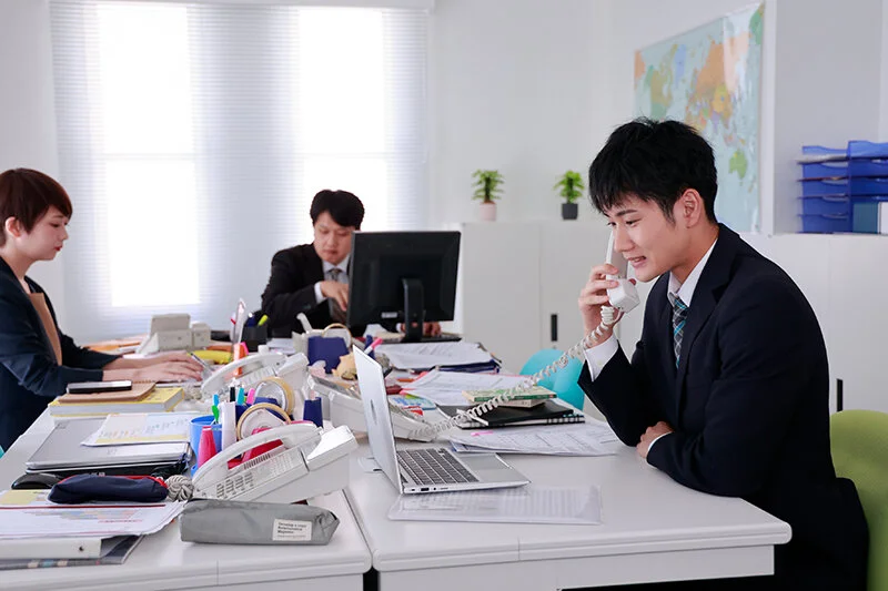オフィスで電話対応をする千歳小梅くん。隣で彩音も仕事に集中している、ふたりのいつもの職場風景