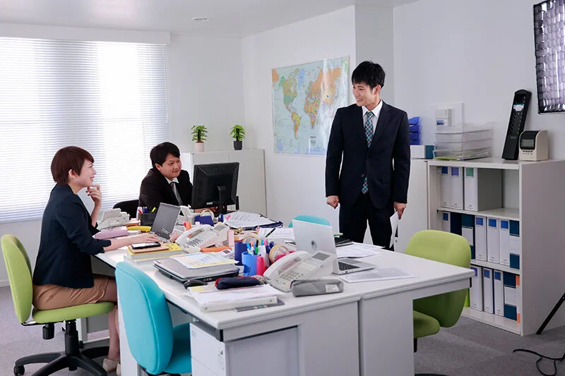 同僚たちと明るく会話する千歳小梅くん。彩音が自然に笑顔を向ける、距離の近い社内の雰囲気